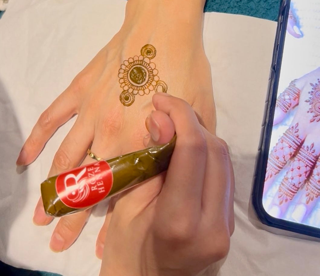 Person holding a Roze Henna cone practising a simple henna design on the back of a hand, with a phone showing henna inspiration nearby